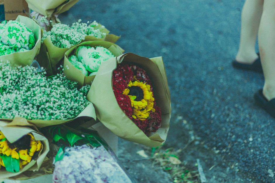 A floral arrangement featuring a bouquet of red and yellow flowers, including a prominent sunflower, wrapped in beige paper. The bouquet is placed on a dark, textured surface, with other bouquets of w