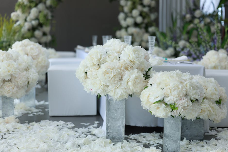 A floral display featuring large, round bouquets of white peonies and carnations arranged in tall, textured concrete vases on a table decorated with scattered white flower petals. The bouquets are lus