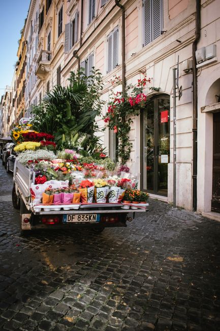 A flower delivery truck parked on a cobblestone street in an urban area with historic European-style buildings featuring cream and beige facades, shuttered windows, and ornate architectural details. T
