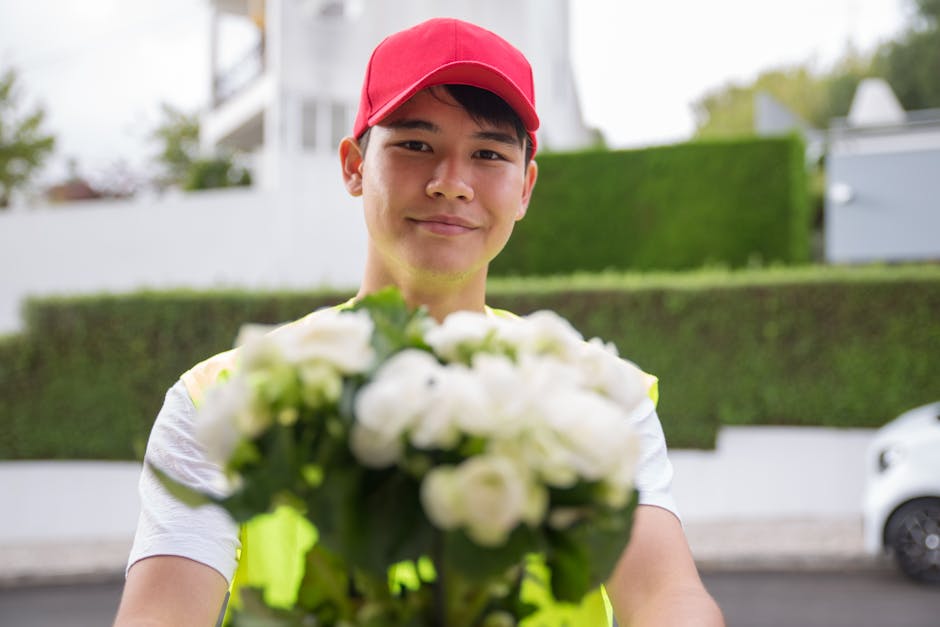 A friendly delivery person wearing a red cap and a high-visibility vest is smiling while holding a bouquet of fresh white roses and green foliage towards the camera. The bouquet is arranged with vibra