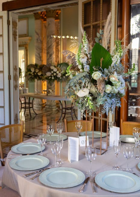 A close-up view of a table setting at a wedding reception, featuring a tall, elegant floral arrangement with white roses, green foliage, pampas grass, and dried elements, displayed on a copper stand. 