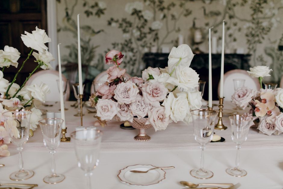 A floral arrangement featuring a large bouquet of white and blush pink roses and lisianthus, displayed in a copper bowl on a wedding table setup. The flowers are fresh, with soft petals and green stem